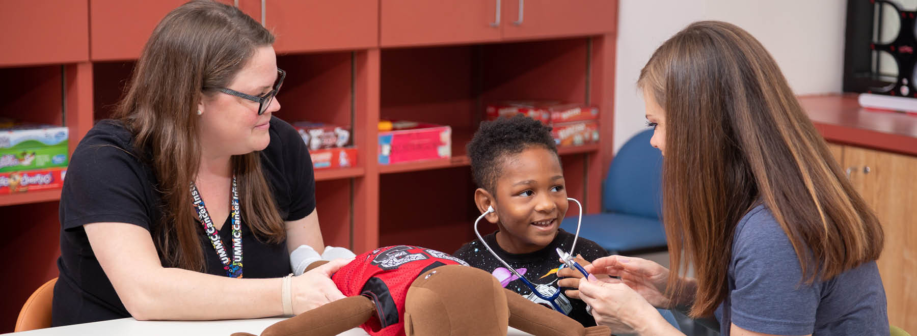 Two child life specialists put a male child at ease by allowing him to use a stethoscope on a stuffed toy.