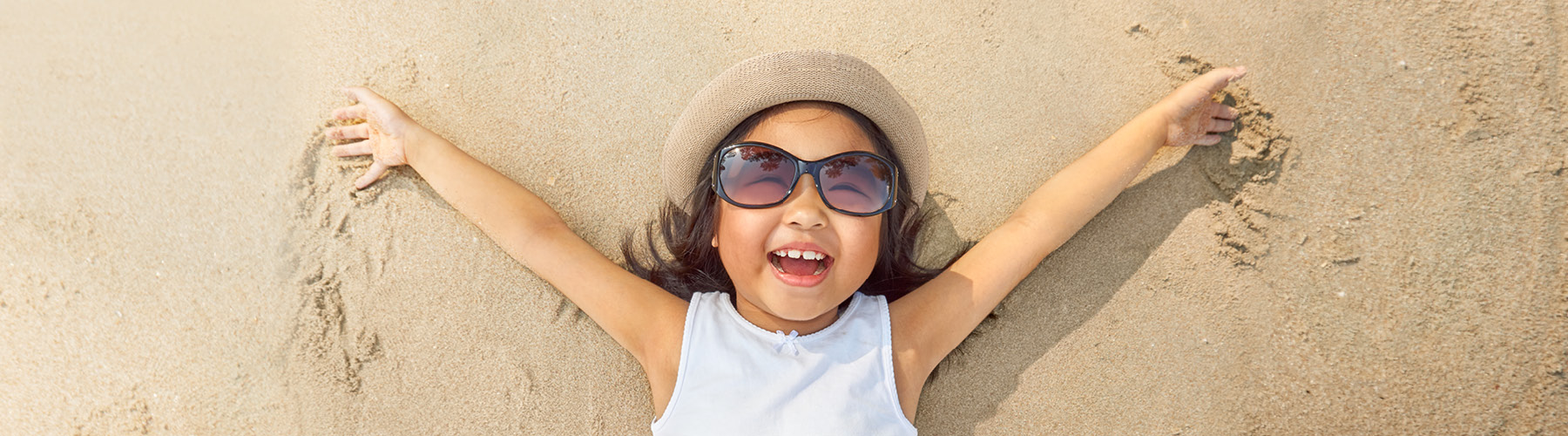 Small girl with sunglasses and hat laying on sandy beach