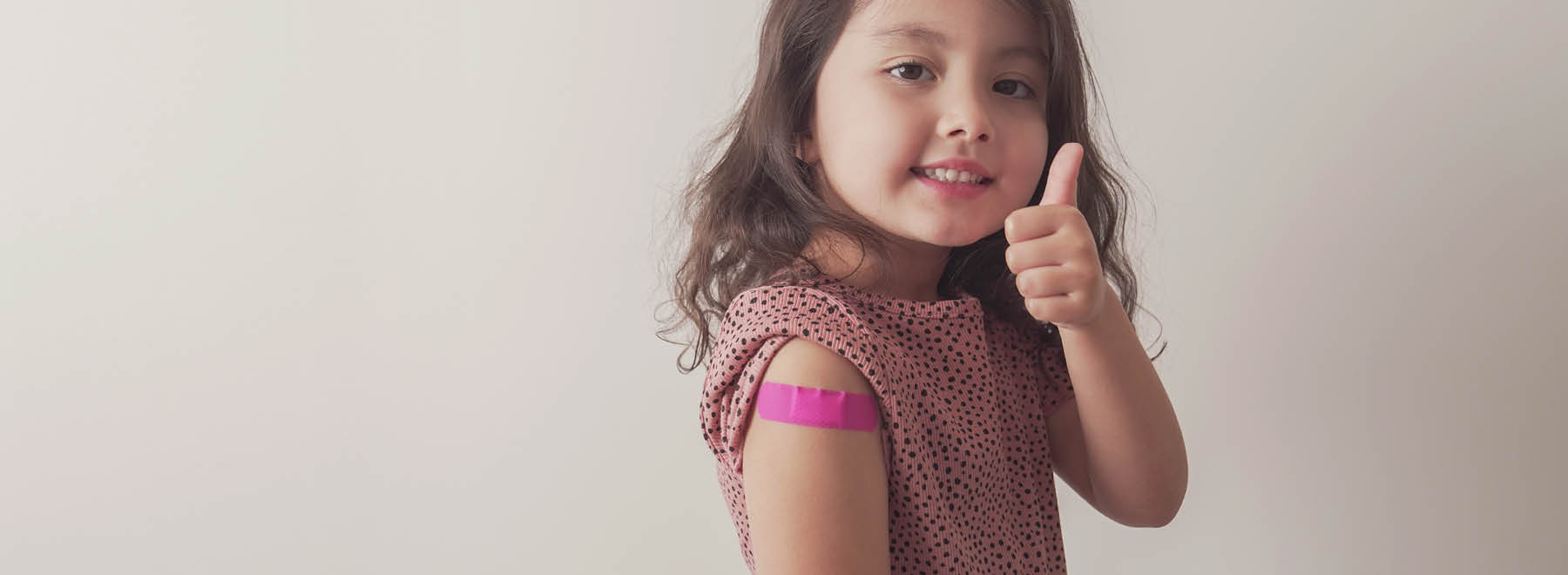A young girl with a bandage on her upper arms gives a thumbs-up.