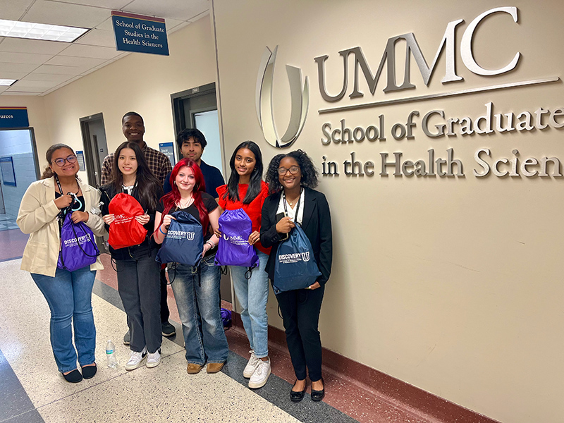Group picture of seven Discovery U students standing outside of the School of Graduats Studies. Five of thm hold branded backpacks.
