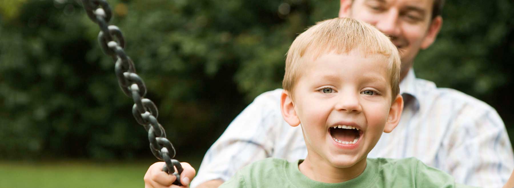 A laughing boy plays on a swing pushed by his father.