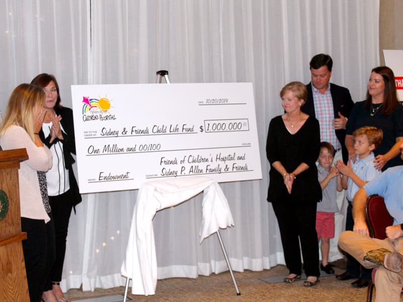 Tiffany Key, left, and Dr. Mary Taylor, second from left, react to the announcment of a $1 million commitment to the Sidney and Friends Child Life Fund while Sidney P. Allen's family members, right, including his wife, Deborah, his children, Sidney Jr. and Rachel, and his grandsons, William and Perry, look on.