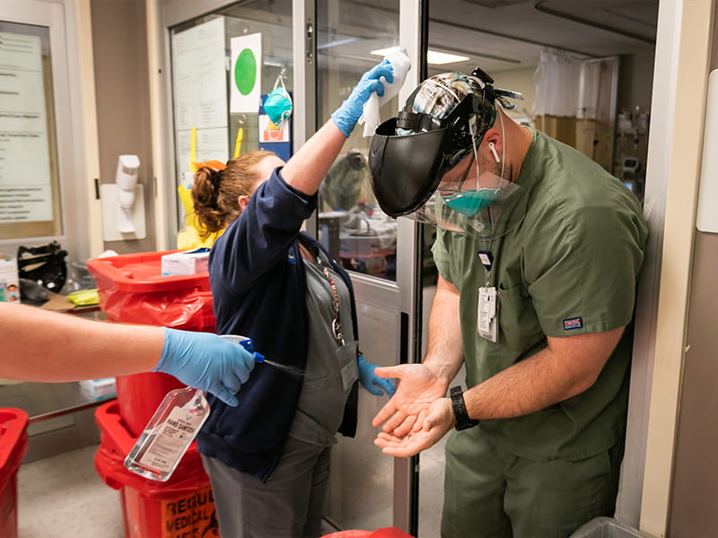 Shannon Moffett, pulmonary nurse practitioner, cleans the face shield of Kevin Marchant, MICU nurse, as another team member sprays his hands with alcohol,