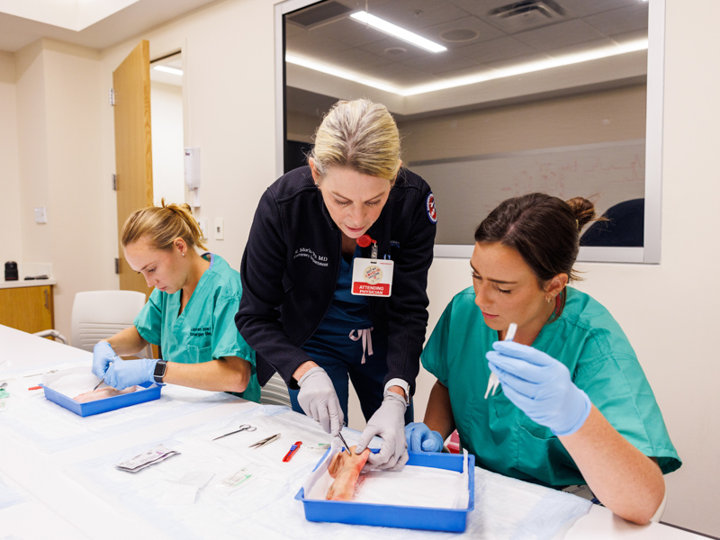 Dr. Risa Moriarty, center, assists new emergency medicine resident, Bethany Fuller, right, with a suturing technique. At work to the left is Lauren Hopper. Jay Ferchaud/ UMMC Communications 