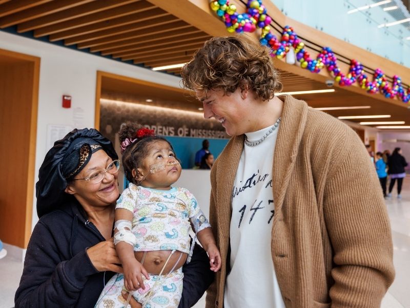 Children's of Mississippi patient LaMiracle Reasor of Waynesboro, held by mom Courtney Reasor, looks up at Ole Miss quarterback Jaxson Dart.