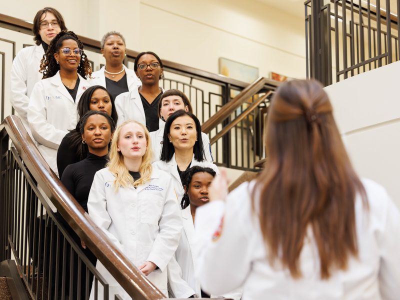 MD-PhD student Madison Hansen, conductor of the Cantus Medici choir, back to camera, directs fellow members during an impromptu rehearsal
