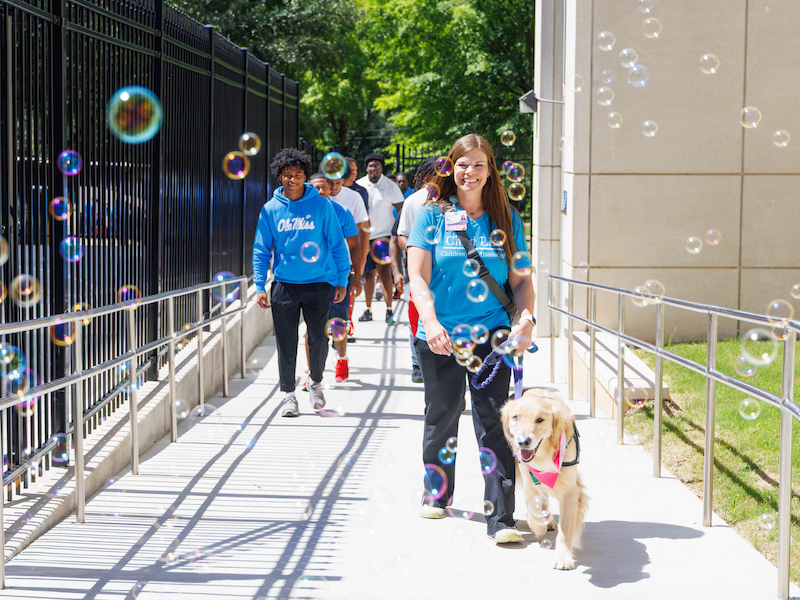Children's child life manager Cara Williams and Hollywood lead the University of Mississippi football team to Mimi's playground.