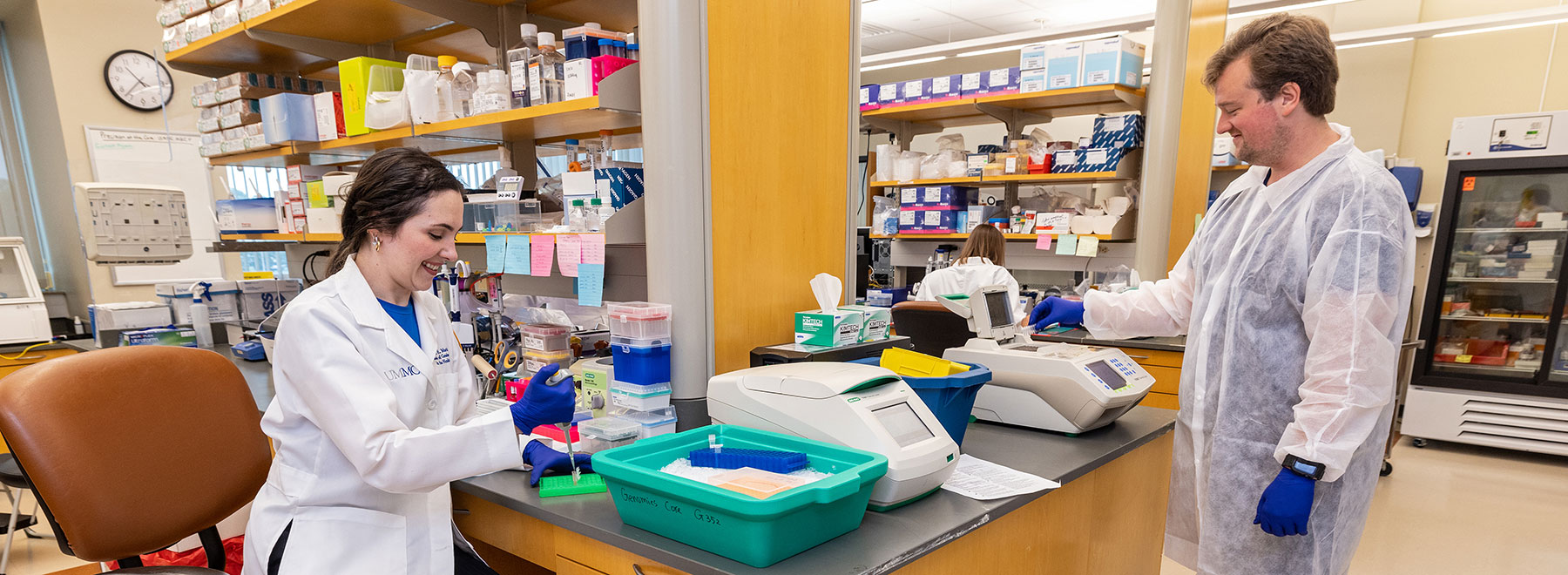 In a research lab, one PhD student add a sample to a vial while another operates a thermal cycler.