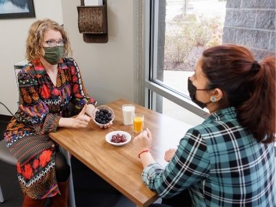 A nutrition counselor, seated at a table with a client, gestures to a bowl of fruit.