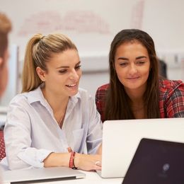 Two women sit together on the same side of a table looking together at a laptop computer screen.