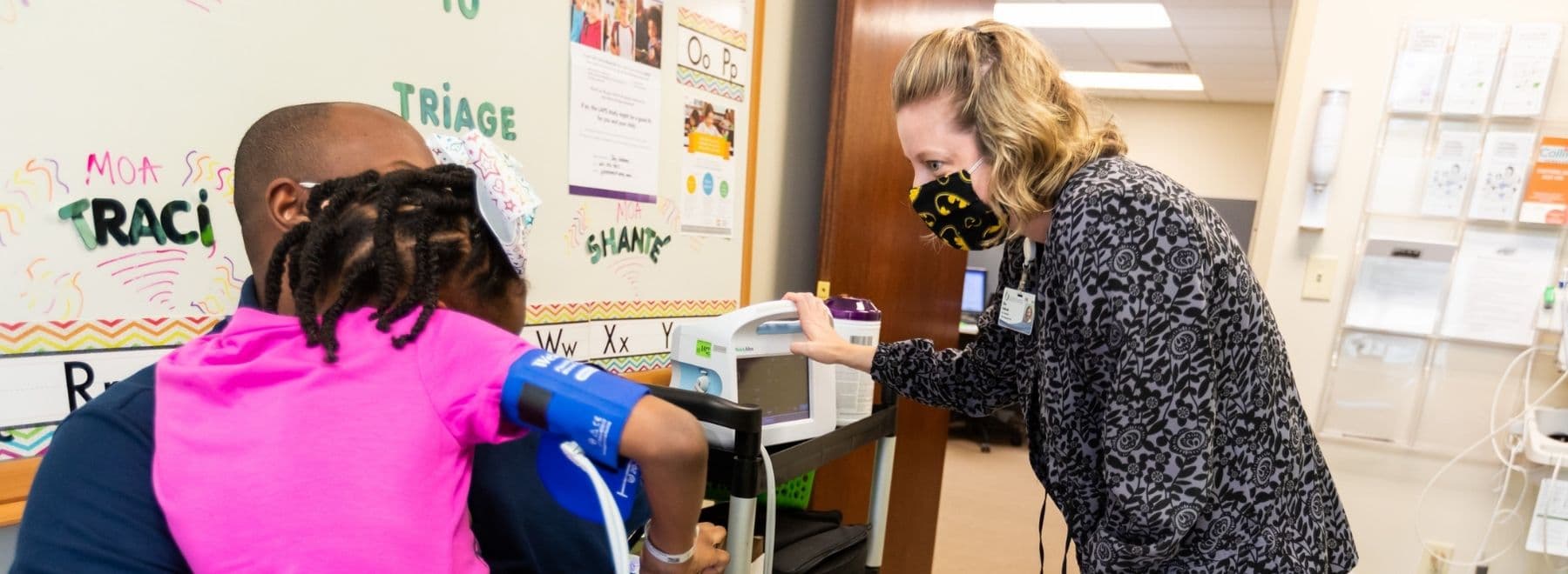 A nurse interacts with a child and parent in a clinical area.