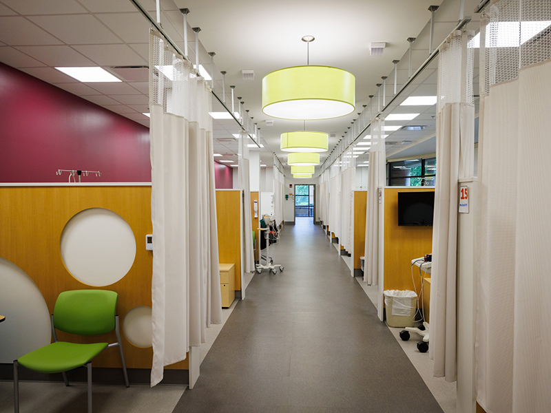 Modern outpatient clinic corridor with curtained exam bays, wood-paneled partitions, lime-green seating, burgundy accent walls, and large circular pendant lights overhead.