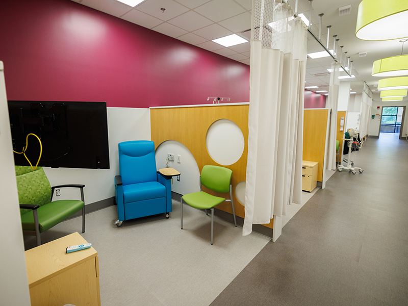 Clinical treatment bay with blue recliner, green guest chairs, privacy curtains, wall-mounted monitor, and wood-paneled partitions with circular accents along an outpatient care corridor.