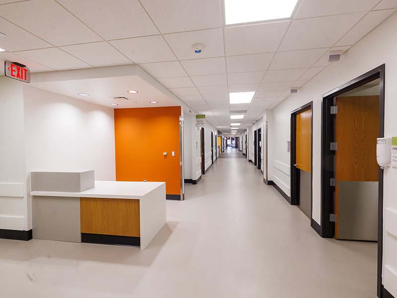 Unit corridor with a white reception desk, orange accent wall, patient room doors and overhead lighting extending down a long hallway.