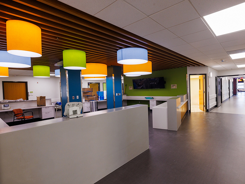 Open nursing station with white work counters, colorful pendant lights, wood-slat ceiling detail, green accent wall, and adjacent patient care corridor.