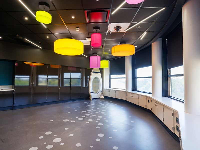 Contemporary activity room with multicolored hanging light fixtures, large windows, built-in counters, dark flooring with circular patterns, and an oval doorway feature.