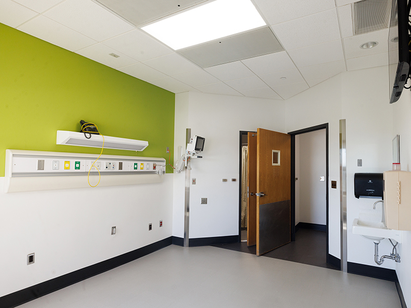 Patient care room featuring a green accent wall, medical headwall unit, sink and storage cabinets, wall-mounted TV, window, and open entry door.