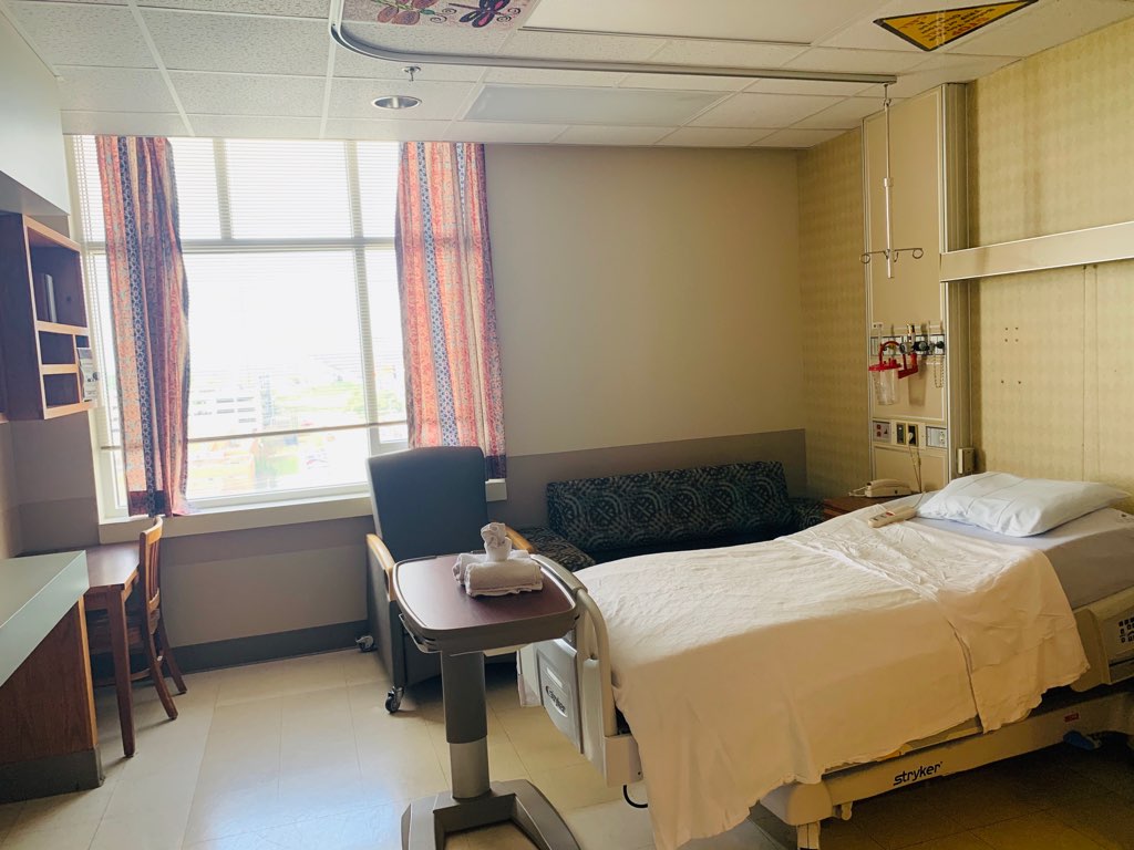 A wide shot of the patient room shows the bed, sofa, chair, desk, cabinets, and rolling try.