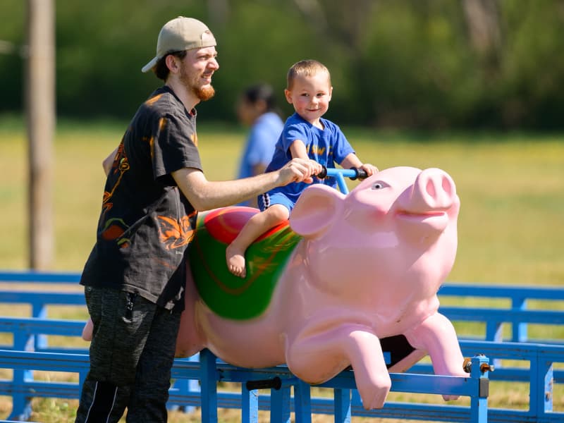 A man in a backward ball cap helps a boy atop a pig ride at Family Fun Day 2026.
