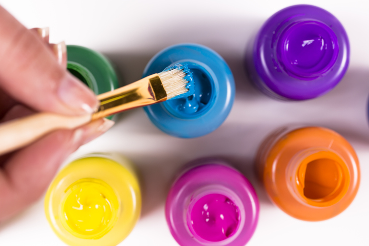 Paintbrush being dipped into blue paint bottle. Multiple colors of paint.