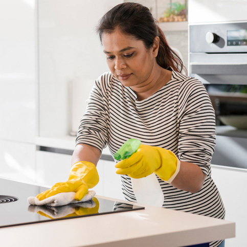 Woman in kitchen wearing rubber gloves while cleaning range with spray bottle of household cleaner and towel.