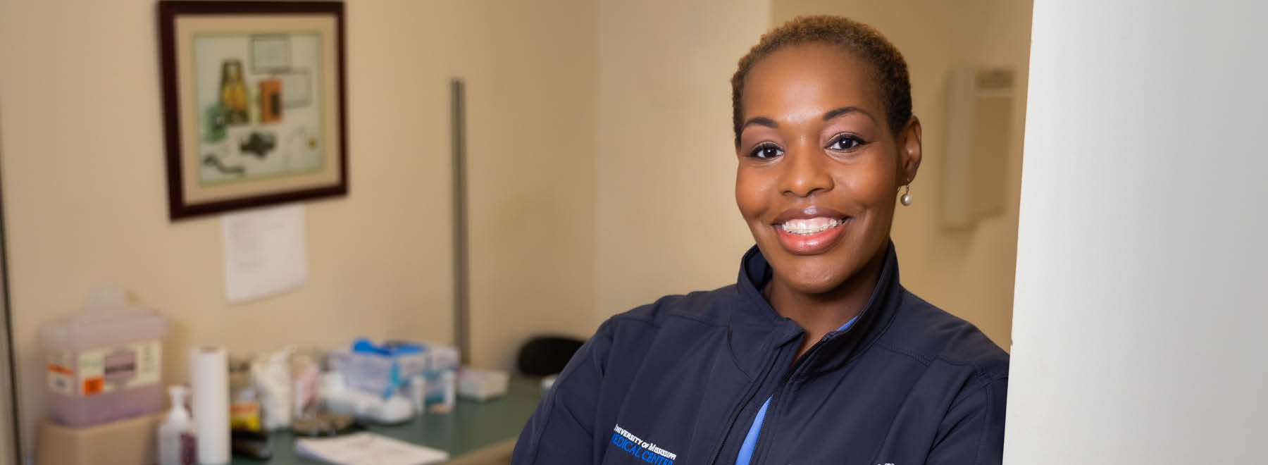 A smiling young woman wearing a UMMC jacket stands leaning against a door frame.