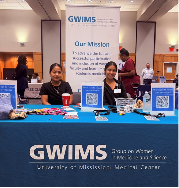 Two GWIMS representatives sit behind a blue information table displaying attendance QR code signs, informational materials, and candy. A large banner behind the table reads, “GWIMS: Group on Women in Medicine and Science, University of Mississippi Medical Center,” and includes the organization’s mission statement. The tablecloth is branded with the GWIMS name and University of Mississippi Medical Center affiliation, while attendees and display booths are visible in the background of a conference or event space.