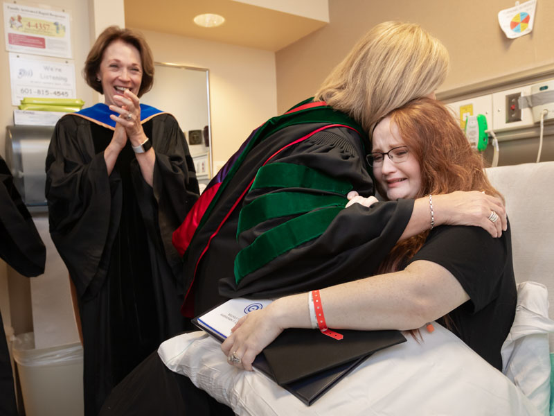 As Dr. Jessica Bailey and others in attendance clap and cheer, Dr. LouAnn Woodward congratulates an emotional Sarah Herrington after presenting her with her diploma in her hospital room.