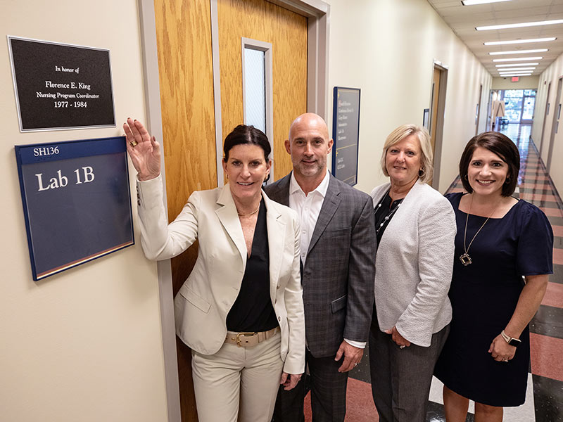 Heather Fennimore of Humanscale, left, proudly points to the plaque outside SHRP Lab 1B indicating the soon-to-be-upgraded student laboratory is dedicated in honor of her aunt, Florence E. King. Joining Fennimore on a tour of the school and the lab’s new location are, from left, Kevin Steets of Humanscale, Kim McGaugh, assistant dean of administration in SHRP, and Dr. Kristi Moore, assistant dean for academic affairs in SHRP.