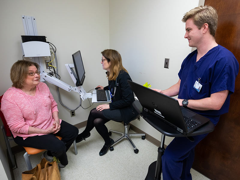 Medical Scribe, in foreground, looks at patient as she answers questions while doctor, in background, looks at a computer.