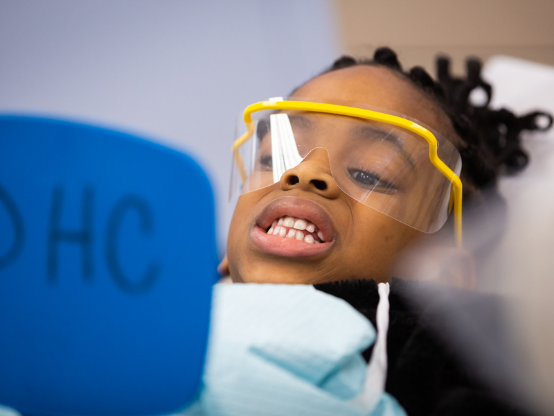 Girl in yellow dental glasses looks at her teeth in mirror.