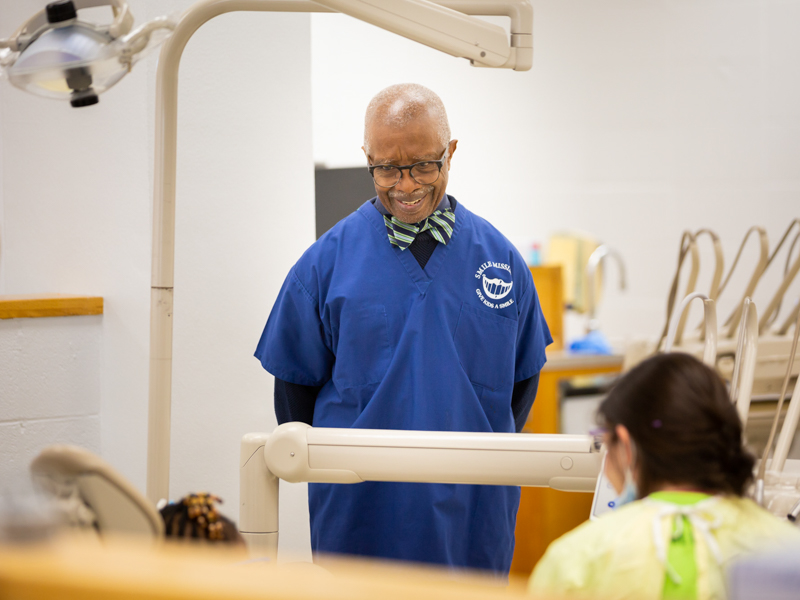 Man in blue scrubs, center, looks at child and dental student in foreground.