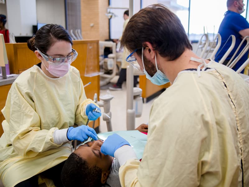 Two dental students work on pediatric patient between them.