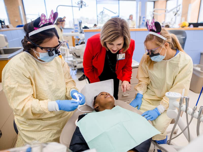 A patient, center, looks up at Dr. Woodward as she chats before two dental students on the left and right begin work.