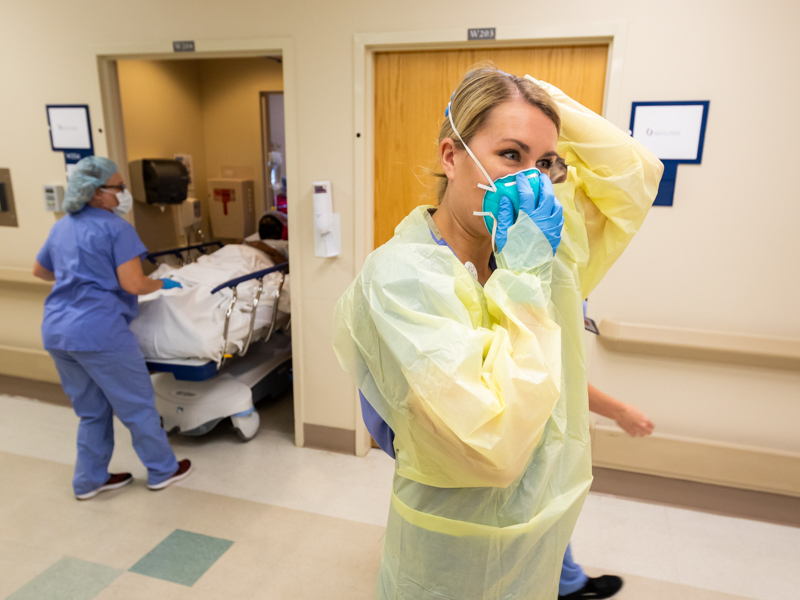 Registered Nurse Amy Brown dons her PPE before entering the room of a COVID-19 positive patient as another patient (background) is moved from her room.