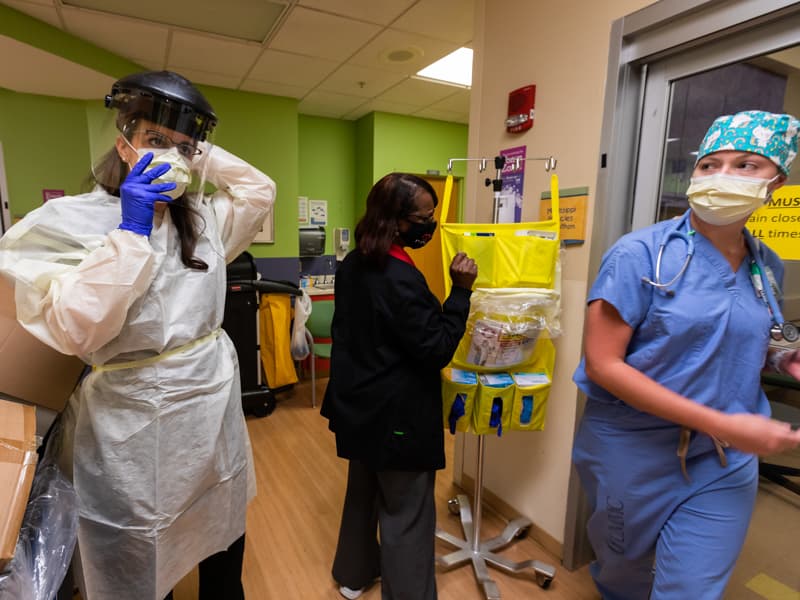 As admissions staffer Arlene Williams, center, and registered nurse Emily Rowell, right, work next to her, Dr. Marlee Wadsworth prepares to enter the room of a suspected COVID patient in the Pediatric Emergency Department.