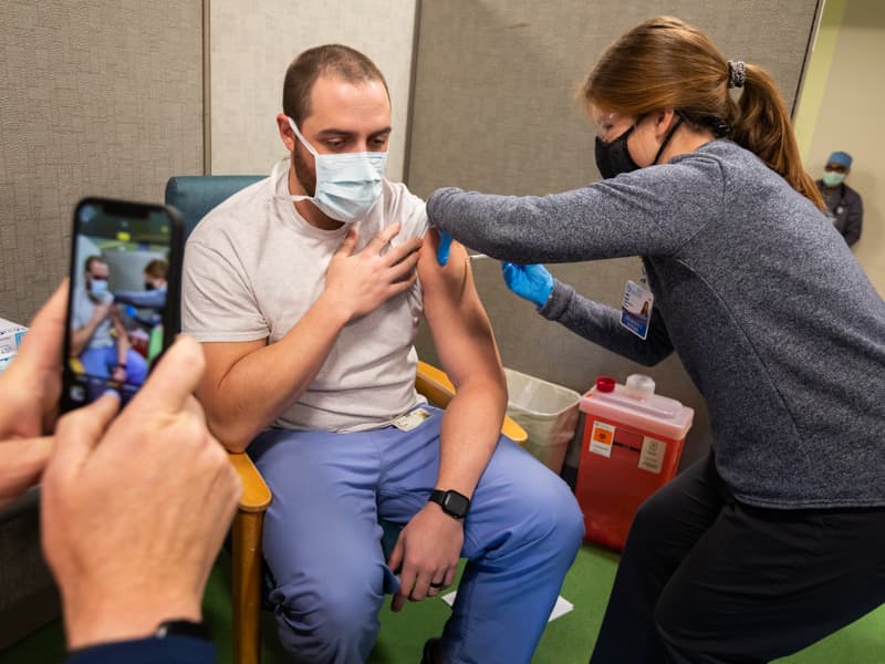 Tyler Fitzgerald, center, a nurse in Antimicrobial Services, receives the COVID-19 vaccine from Kaitlin Gaskin, pharmacy intern, while Carl Mangum, associate professor of nursing, captures video for Fitgerald to share with his wife.