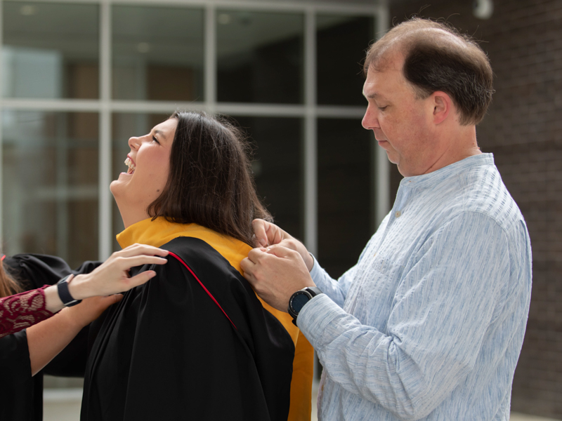 Dr. Christopher Jackson helps his daughter, Kimberly Jackson, with her hood alongside fellow School of Graduate Studies in the Health Sciences friend Sydney Thomas prior to UMMC's commencement ceremonies May 27 at the Mississippi Coliseum. Jackson graduated from UMMC in 1998, the year his daughter was born.