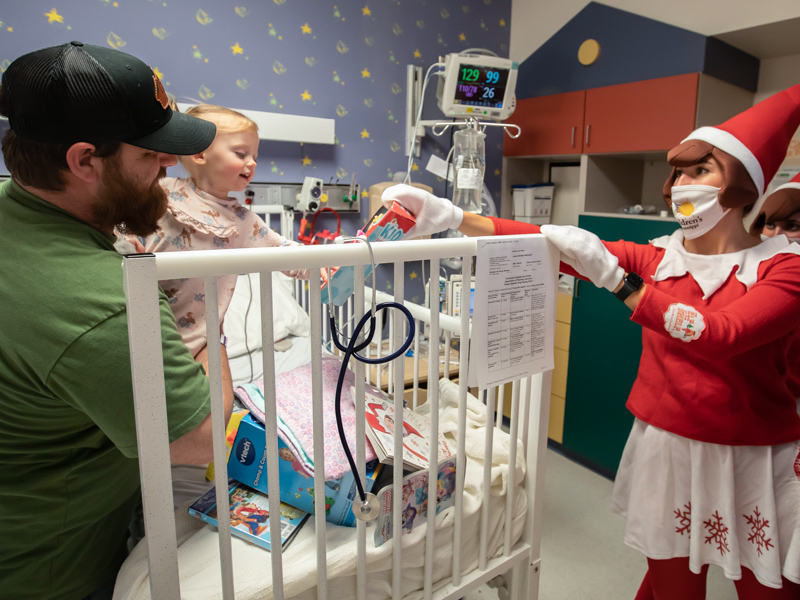 Children’s of Mississippi patient Margaret Booker is held by her father, Brandon Booker, as one of Santa Claus' elves, Cillie Taylor, gives her a doll during their inpatient visit at Blair E.Batson Tower Wednesday.