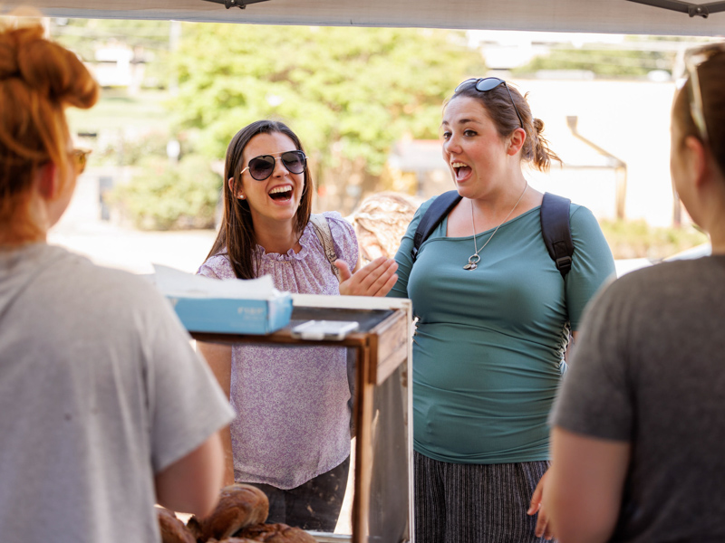 Lauren Greenlee, left, and Nicole Connolly, both admissions officers with Student Services and Records, shop the Sunflower Oven co-op bakery stand at Thursday afternoon's Farmer's Market behind Backyard Burger. The event was sponsored by the UMMC Office of Well-being. Joe Ellis/ UMMC Communications