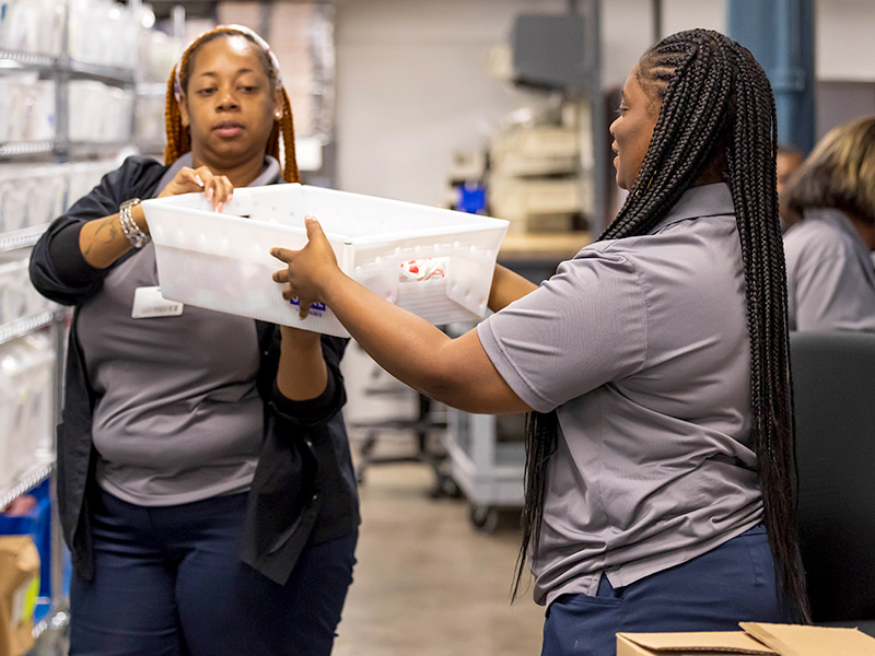 Supply techs Diedre Hobson, left, and Adreyonna Kyser work together to pass off bins and fill orders in the Supply Chain Department.