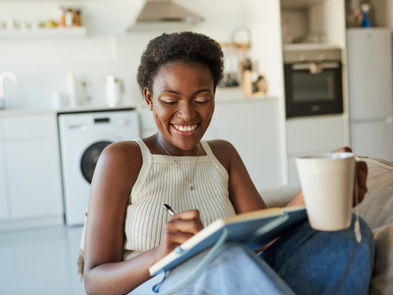 Shot of a young woman having tea and writing in a notebook on the sofa at home.