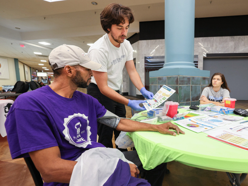 Medical student Shade Smith, standing, describes the Healthy Eating Plate to Ernest Galloway of Vicksburg while monitoring his blood pressure during a November 5 health screening at the Vicksburg Mall. Medical student Rachael Todd, right, is also part of the Student Health Coalition team. Jay Ferchaud/ UMMC Communications 