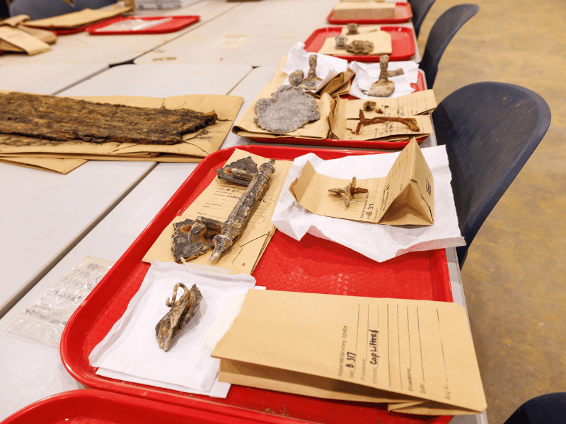 Objects found in Asylum Hill graves include a cap lifter (left, foreground) shaped like a calla lily and used to lift or replace a wooden panel that covered a glass viewing pane set in the coffin lid; and, beside it, a coffin handle. To the right of the coffin handle is a thumbscrew and escutcheon set; these were used to hold the coffin lid closed, as opposed to nailing it down.