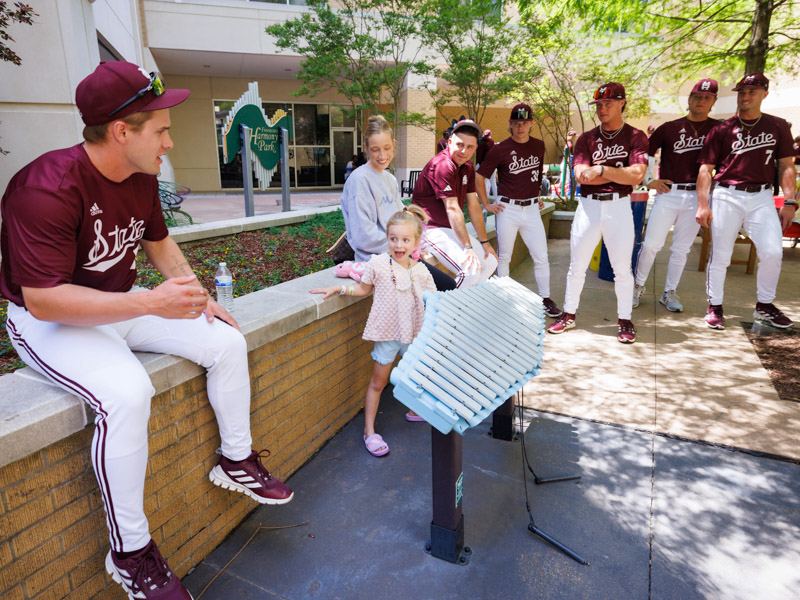 Laurel Mathis, of Brandon, a patient at Children's of Mississippi, reacts to MSU infielder/outfielder Aaron Downs as her mother, Kyla Mathis, and Bulldog players Nate Chester, Bryce Chance, Cam Schuelke, Hunter Hines and Connor Hujsak, look on.