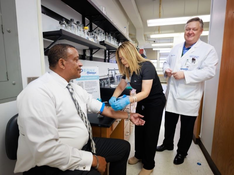 Yelena Neely, center, a research associate with the Department of Pharmacology and Toxicology, draws blood from Kevin Wilbert, director of operations for centers and cores in the department, with encouragement from Dr. Ken Butler, professor of pharmacology and toxicology and associate director of the UMMC Biobank.