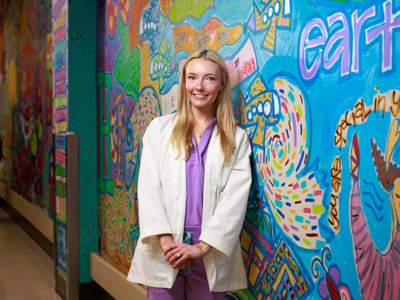 Medical student and artist Virginia Kate Gammon, editor of the arts and literature journal, The Mosaic, draws on the lessons she learned from her late grandmother, artist Virginia Thomas, who gave free art lessons to children. Gammon is shown here, appropriately, beside artwork displayed in the hallway leading to the Batson Tower of Children’s of Mississippi.