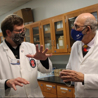 Two men wearing white coats and masks (Dr. Griggs and Dr. Mecholsky) discuss materials science.