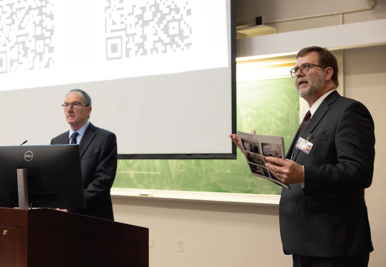 Dr. Richard Price standing at a podium. Dr. Jason Griggs is to his right speaking.