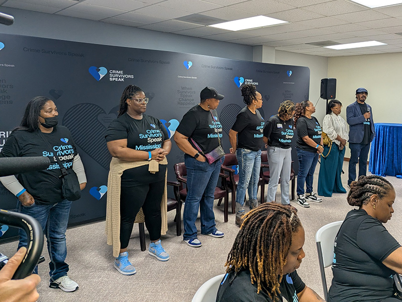 Group of eight participants standing in a line wearing Crime Survivors Speak Mississippi shirts. The last individual has a microphone.